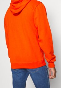 Sudadera con capucha naranja de corte regular, con puños y dobladillo acanalados. La tela parece suave y se complementa con una capucha abierta y relajada.