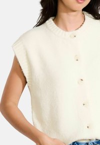 Krēmveida vilnas maisījuma vestes ar ribotu apkakli, īsām piedurknēm un pieciem priekšējiem pogām. Mīksta tekstūra, ikdienišķs dizains, ideāli piemērots slāņošanai.