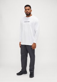 Hosszú ujjú fehér póló, a mellkason "Calvin Klein" logóval, sötétszürke nadrággal és fekete sportcipővel párosítva. Kényelmes szabás és lezser stílus.