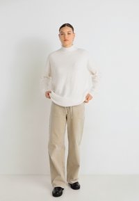 Lengvas baltas golfas su šonkaulinio rašto tekstūra, derinamas su smėlio spalvos platėjanciais kelnėmis ir juodais batais, stovintis prieš baltą foną.