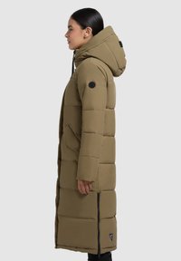 Cappotto lungo e voluminoso di colore verde oliva con cappuccio, dotato di zip frontale, tasche laterali e toppe con logo sulla manica; texture liscia, design fino al ginocchio.