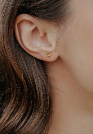 Gold smiley face stud earring in a pierced ear, with smooth round shape and simple facial features. Brown hair partially visible.