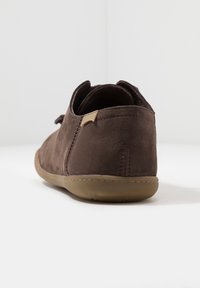 Zapato de ante marrón con un perfil bajo, puntera redondeada y suela de goma suave. Presenta un diseño cosido y una pequeña lengüeta trasera para facilitar el uso.