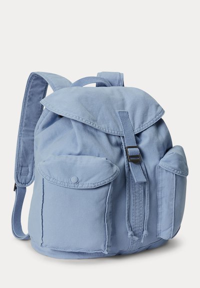 Mochila de tela azul claro con dos bolsillos frontales con botones, tapa con solapa asegurada por correa y hebilla, y correas acolchadas para los hombros.