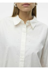 Camisa blanca de botones con cuello, que presenta pequeños adornos de plata dispersos. Textura de tejido suave, ajuste entallado alrededor del cuello.