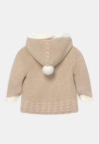 Beige stickad babyjacka med huva som har en vit fluffig pom-pom, kabelstickade mönster och vit fleecekanter på ärmslut och huvan.