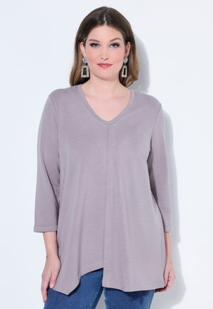 Kvinde iført lys lilla V-hals langærmet top med asymmetrisk kant, blå jeans og store geometriske sølvøreringe, stående mod en hvid væg.