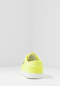 Zapatilla de lona amarilla neón con suela de goma blanca, que presenta un diseño minimalista y costuras acentuadas en los laterales.