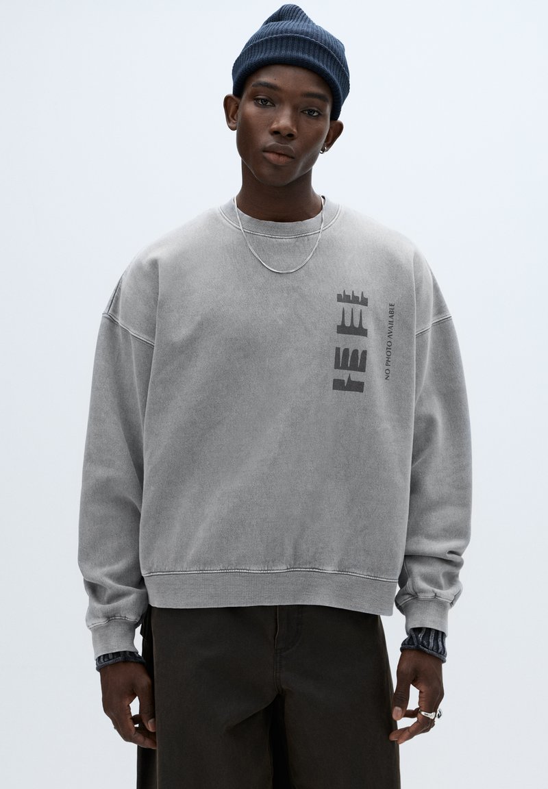 Harmaa ylivuotoinen collegepaita, jossa on teksturoitu pinta ja pyöreä pääntie; siinä on musta graafinen kuvio sekä resorit helmassa ja hihansuuissa.