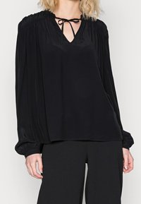 Black blouse with a V-neck and tied front, featuring gathered shoulders and long, billowing sleeves. Smooth, shiny fabric.