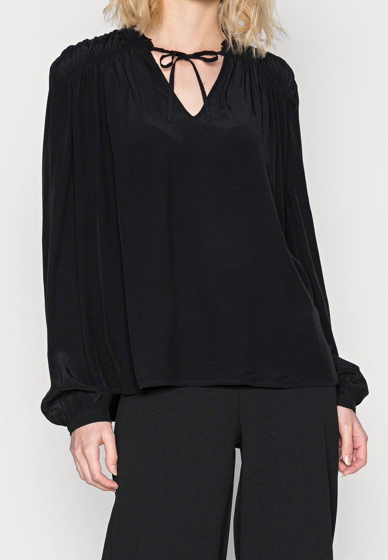 Black blouse with a V-neck and tied front, featuring gathered shoulders and long, billowing sleeves. Smooth, shiny fabric.
