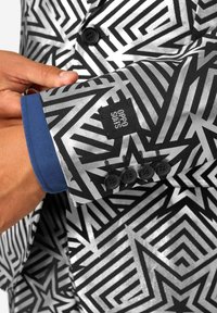 Chaqueta de traje negra y plateada con patrones geométricos, que presenta un puño azul, botones negros y una etiqueta de marca en la manga.