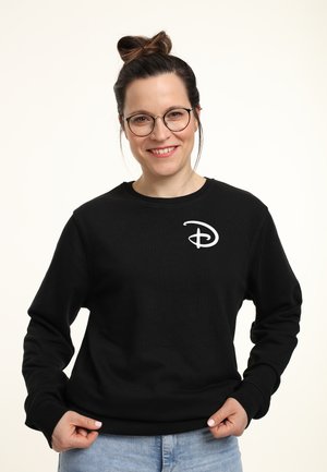 Smilende kvinde med briller og hår i knold iført sort sweatshirt med hvid Disney-logo, holder nederdelen, står mod en ensfarvet baggrund.