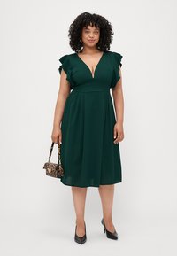 Robe verte foncée avec des manches courtes à volants, un décolleté en V profond et une jupe évasée fluide, associée à un sac à main imprimé léopard et des talons noirs.