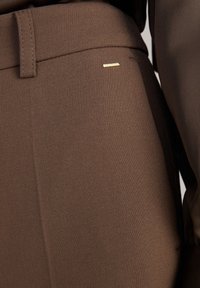 Tissu marron texturé en gros plan de pantalons sur mesure avec ceinture, passant de ceinture et petit détail rectangulaire doré près de la poche.