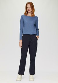 Langsleeve blauwe top met een vierkante hals, gecombineerd met zwarte cargo broek met zijzakken. Model draagt witte sneakers.