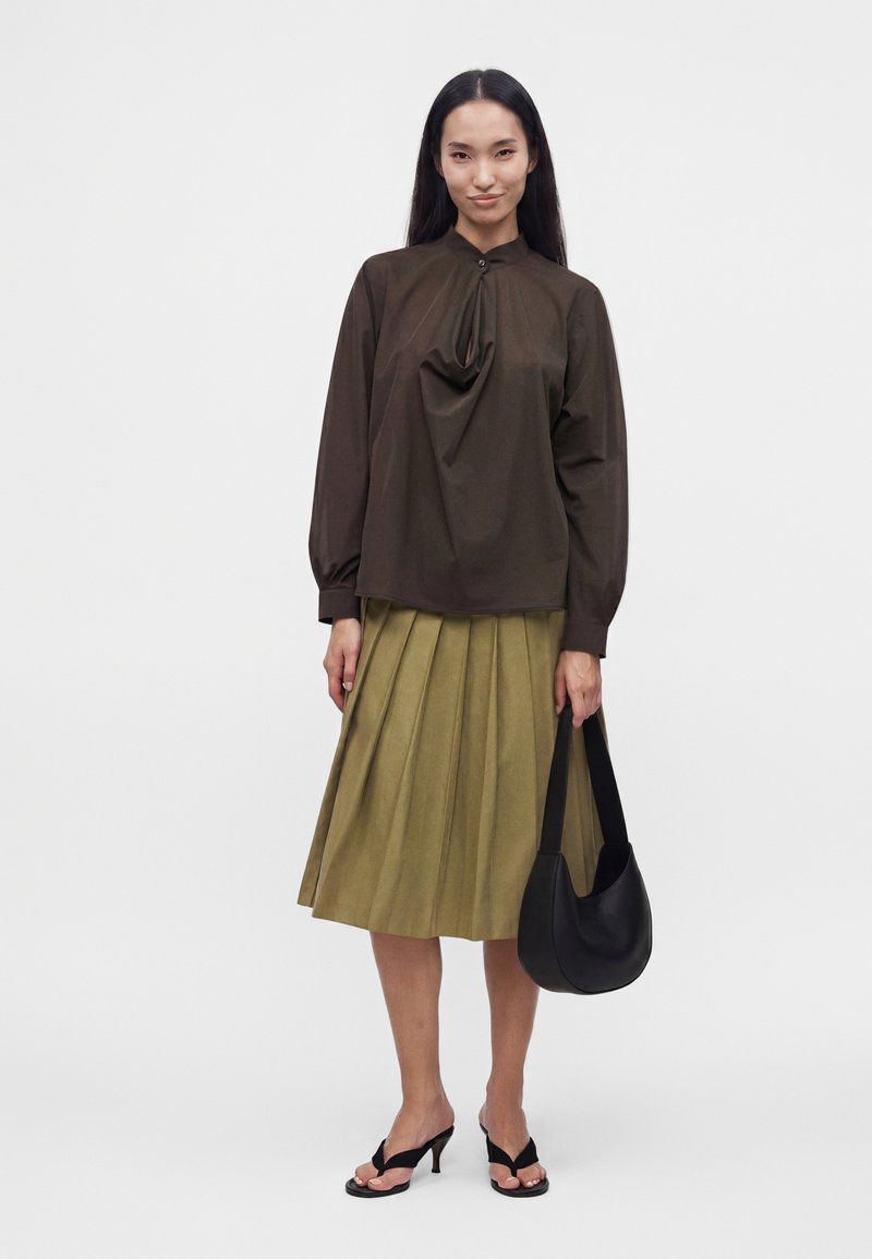 Femme portant un chemisier à manches longues marron foncé, une jupe plissée vert olive, des sandales à talons noires et tenant un sac à main noir.