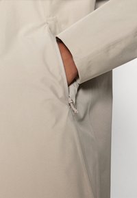 Lys beige frakk med lomme på siden med glidelås, subtile sømdetaljer og en glatt tekstur. Designet er minimalistisk og funksjonelt.