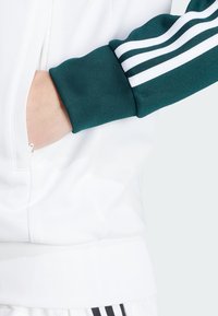 Bílá mikina na zip s tyrkysovými rukávy, zdobená třemi bílými pruhy. Rukávy jsou ribbené a látka vypadá hladce a sportovně.