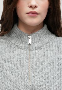 Γκρι πλεκτός μαζεμένος με ψηλό γιακά και μισό φερμουάρ. Διαθέτει υφασμάτινη ριβέ ραφή και ασημένιες λεπτομέρειες για το φερμουάρ.