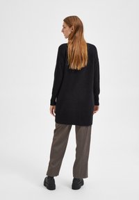 Un long pull noir avec une coupe décontractée, doté d'un ourlet asymétrique, de poignets côtelés, porté sur un pantalon rayé gris et des chaussures noires épaisses.