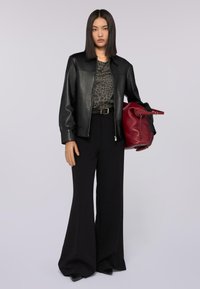 Czarna skórzana kurtka na wzorzystej bluzce, połączona z szerokimi czarnymi spodniami. Trzyma czerwoną torbę, włosy proste i ciemne.