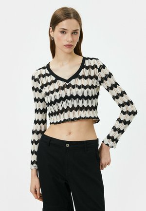 Schwarzes und weißes gehäkeltes Crop-Top mit V-Ausschnitt, das ein gewelltes Streifenmuster und lange Ärmel zeigt, getragen über schwarzen Hosen.