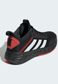 Zapatilla deportiva negra con parte superior de malla, acentos blancos a rayas, detalle rojo en el talón y suela negra texturizada. Sistema de cordones en la parte frontal.