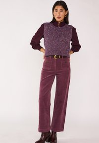 Gilet in maglia viola con macchie multicolori sopra una camicetta bordeaux scuro, abbinato a pantaloni a gamba larga in velluto a coste bordeaux e stivaletti con tacco.
