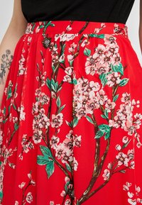 Jupe rouge à fleurs avec un motif de cerisiers en fleurs roses et de feuilles vertes, au design plissé, avec une taille ajustée et une texture de tissu lisse.