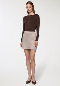 Brązowa bluzka z długim rękawem zestawiona z beżową zamszową mini spódnicą z przodu z rozcięciem. Model ma na sobie brązowe skórzane klapki na obcasie; widoczne są gładkie faktury.