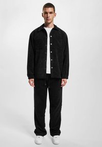 Black corduroy jacket with snap buttons, paired with matching pants. Model wears a white t-shirt and white sneakers.