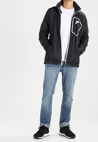 Svart lättviktsjacka med hög krage, vit dragkedja och en vit logotyp på bröstet. Bärs över en vit t-shirt och denimjeans.