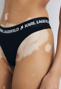 Black thong underwear with a wide elastic waistband featuring "KARL LAGERFELD" logo in white. Smooth material, minimalist design, fitted shape.