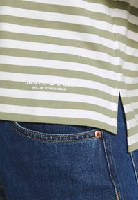 Stribet grøn og hvid bomulds t-shirt med sideslidser og logodetalje. Båret over blå denim jeans med kobberknapdetaljer.