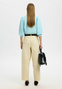 Světle modrá blůza s nafouknutými rukávy, béžové široké kalhoty, černý pásek, držící malou černou tašku s připevněným vzorovaným šátkem.