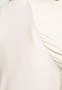 Camiseta de un beige claro con una textura de tela suave, que presenta detalles fruncidos en el hombro y un escote simple y redondeado.