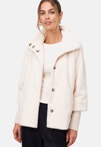 Veste en fausse fourrure crème avec un large col, quatre boutons brillants et des manches trois-quarts. Texture douce avec une coupe décontractée.