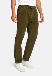 Pantalones de pana verde oliva con pliegues frontales, diseño ajustado y un acabado texturizado, combinados con zapatillas blancas.