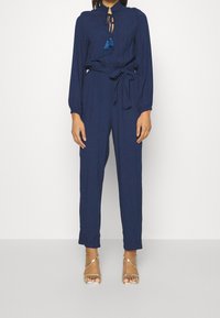 Navy blue jumpsuit with long sleeves, tie neckline, textured fabric, and a belted waist; features decorative blue tassels. Worn with gold sandals.