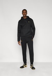 Μαύρο φούτερ με κουκούλα και τσέπη καγκουρό, συνδυασμένο με μαύρο φόρμα με πορτοκαλί κείμενο "UFC", ολοκληρωμένο με μαύρα και μπεζ αθλητικά παπούτσια.