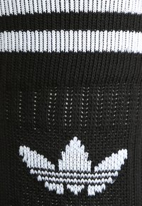 Crni pleteni materijal s bijelim horizontalnim prugama i bijelim izvezenim troklanim logotipom na dnu. Tekstura izgleda mekana i fleksibilna.