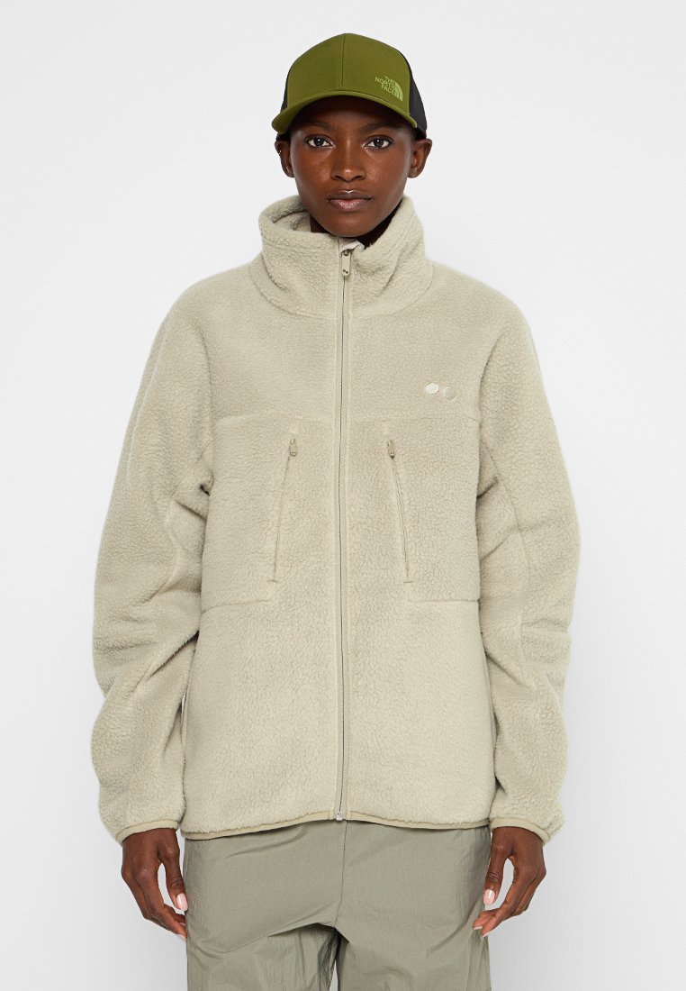 Hellbeige Fleecejacke mit hohem Kragen, zwei vorderen Reißverschlusstaschen und einer strukturierten Oberfläche. Kombiniert mit einer grünen Mütze.