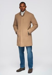 Beige Wollmischungsmantel mit Stehkragen, seitlichen Taschen und geradem Schnitt. Getragen über einem hellen Rollkragenpullover und kombiniert mit blauen Jeans und braunen Stiefeln.
