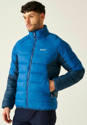 Mężczyzna w niebieskiej kurtce puchowej Regatta z kieszeniami na zamek, stojący z jedną ręką w kieszeni, patrzący w lewo na tle jednolitego tła.