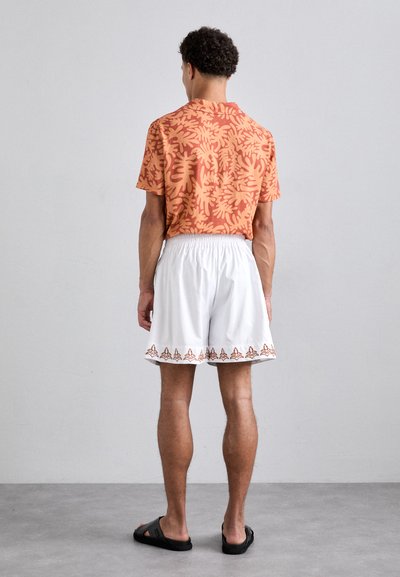 Short-sleeve orange shirt with a leafy pattern, paired with white shorts featuring a decorative hem, worn with black sandals.