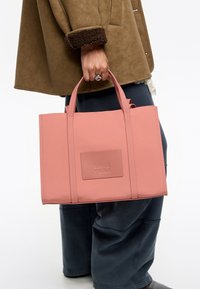 Różowa torba na ramię wykonana z materiału syntetycznego, o prostokątnym kształcie, z podwójnymi uchwytami i wypukłą naszywką z logo na przodzie.