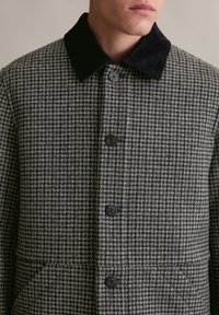 Houndstooth patroonjas gemaakt van een wolmix stof, met een zwarte corduroy kraag en vijf zwarte knopen, voorzien van twee voorzakken.