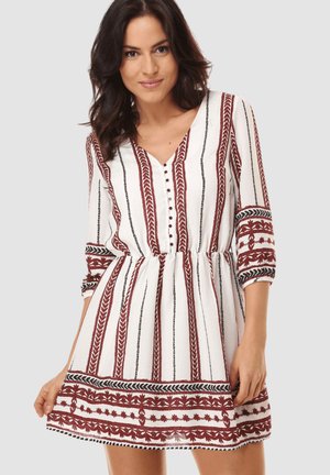 Mujer con vestido blanco con rayas verticales bordadas en granate y negro, mangas tres cuartos, botones en la parte delantera y cintura fruncida.