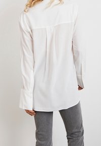 Long-sleeve, white blouse with a back pleat, buttoned cuffs, and a curved hem, made from light, smooth fabric.